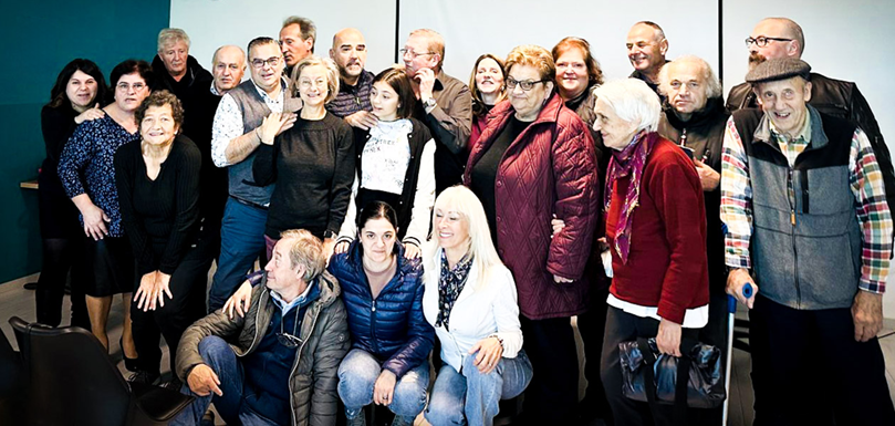 Eine größere Gruppe von Menschen posiert zusammen in einem Raum vor einer weißen Wand mit zwei Leinwänden. Alle lächeln und stehen eng beisammen.